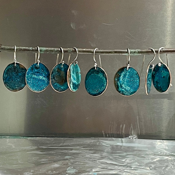 Set of copper with blue patina  earrings hanging on a rod against a reflective surface.